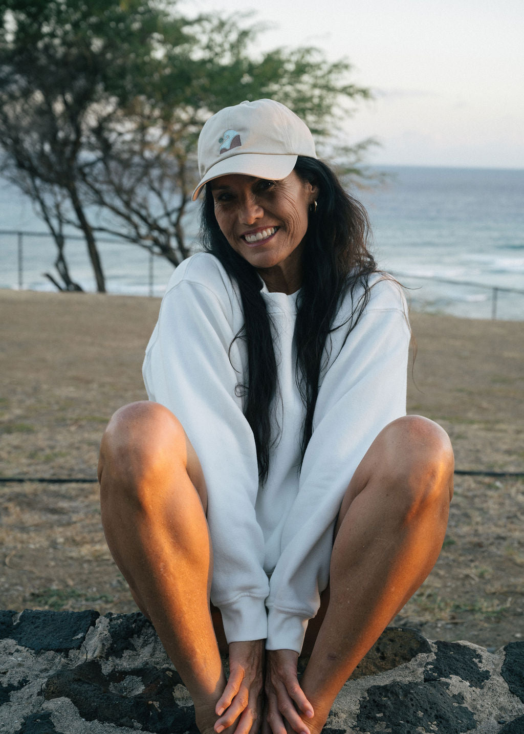 Woman sitting in park near ocean wearing Coastal Range baseball cap and white sweatshirt.