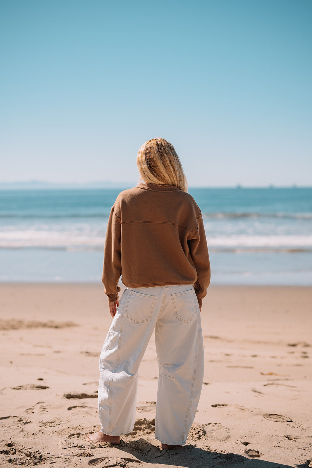 Person wearing white pants and brown sweatshirt looking at ocean