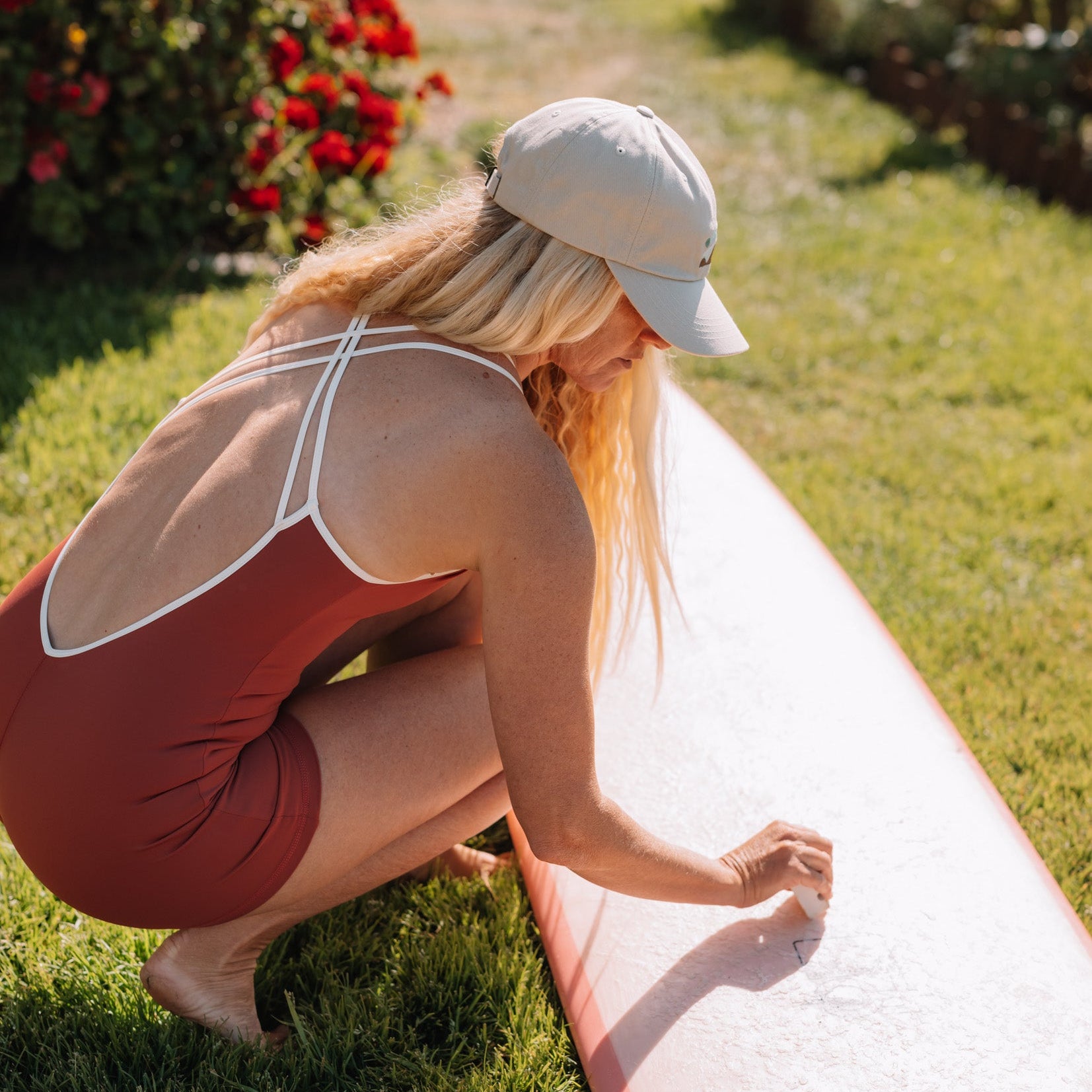 Woman in a red dswimsuit and beige cap waxing a surfboard.