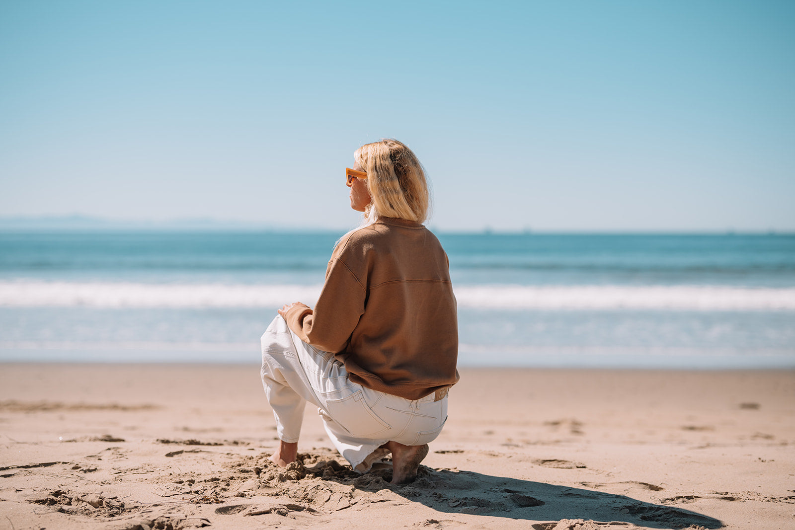 Person wearing brown sweatshirt and sitting on a beach looking at the ocean