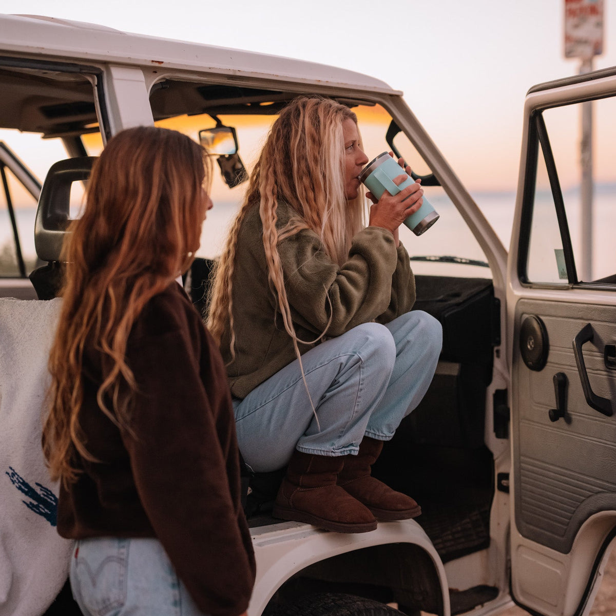 Two women sitting in the open door of a white vehicle at sunset, with one drinking from a blue tumbler. The scene is outdoors near a body of water, with warm golden hour lighting.