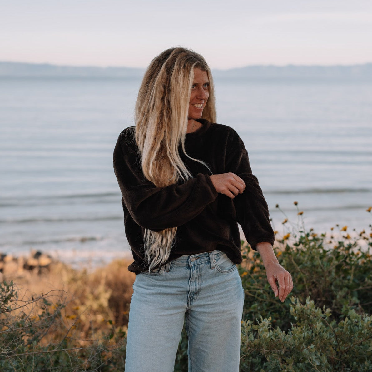 A woman stands outdoors near the ocean wearing a brown long-sleeve top and light blue jeans, with shrubs in the foreground and a calm sea under a pale sky behind her.