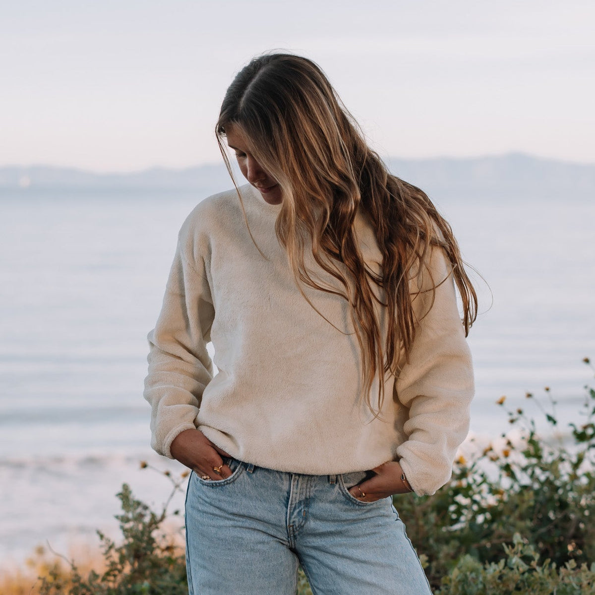 Person wearing a beige sweater and light blue jeans standing by a body of water.