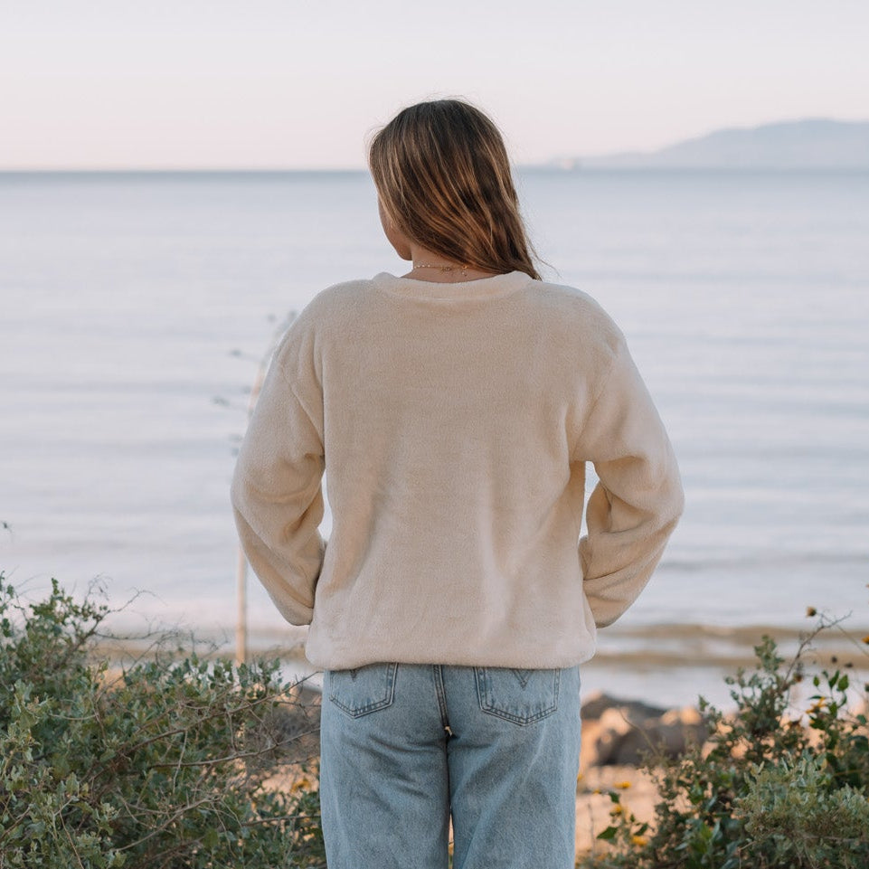 A woman stands with her back to the camera, looking out over a calm sea at sunset. She is wearing a cream-colored long-sleeve knit sweater and light blue wide-leg jeans, positioned on a dirt path surrounded by green coastal shrubs.