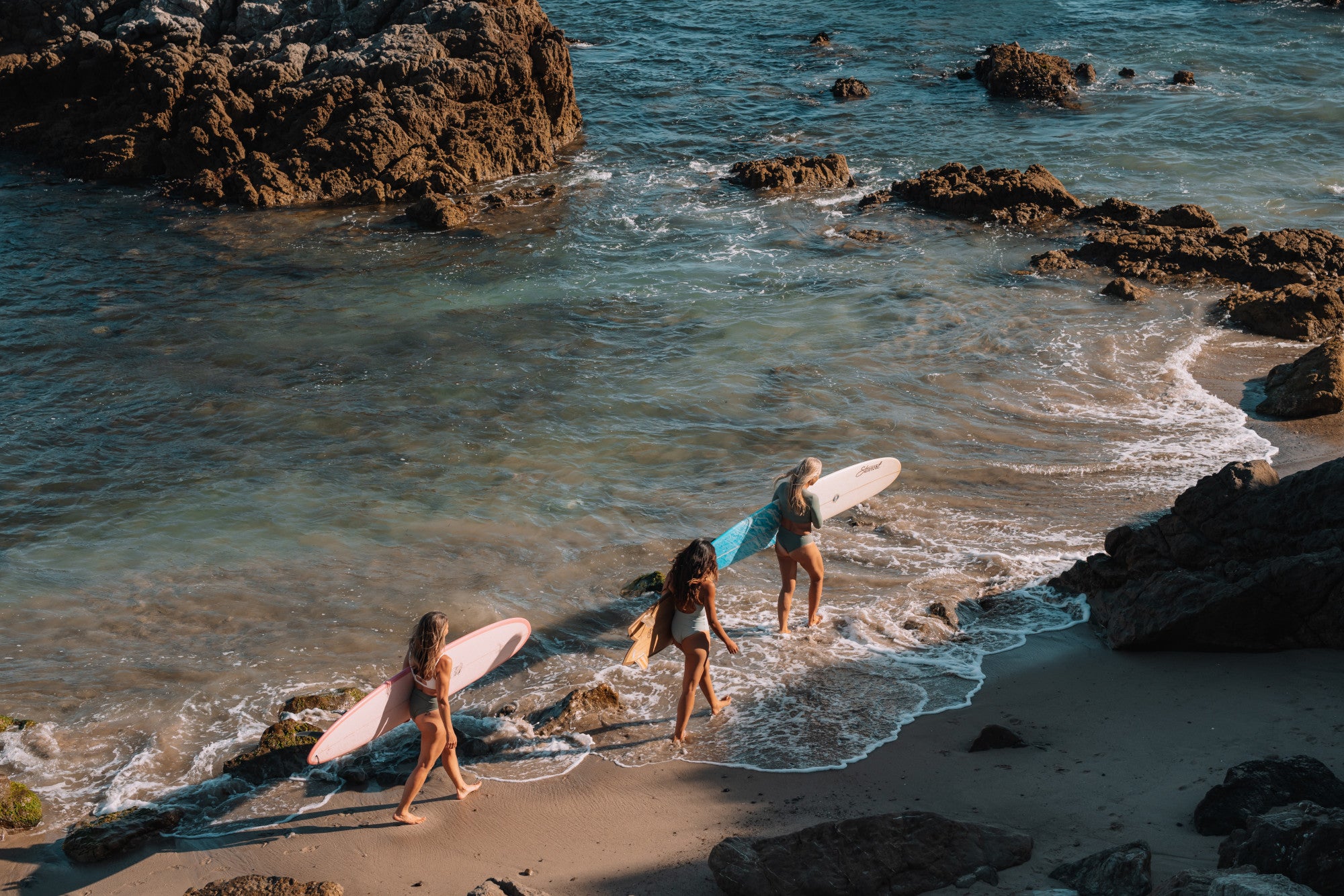 Three surfers with boards walking along a rocky beach.