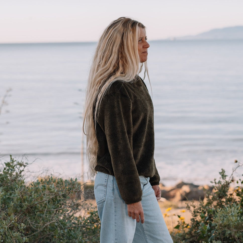 A woman stands barefoot on a beach path, wearing a dark olive green long-sleeve top and light blue wide-leg jeans, with ocean and shrubs in the background.