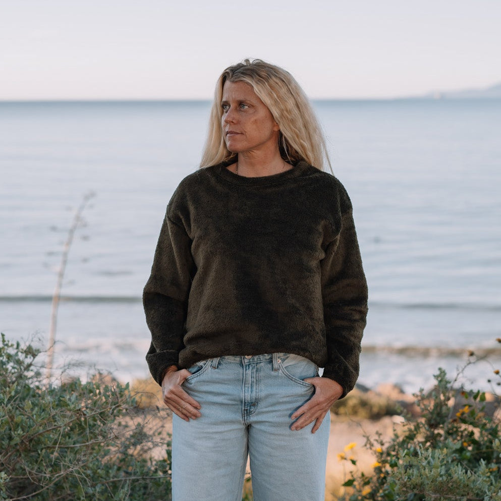 A woman stands barefoot outdoors by the water, wearing a dark olive-brown long-sleeve top and light-wash blue jeans, with shrubs in the foreground and a calm sea under a pale sky behind her.