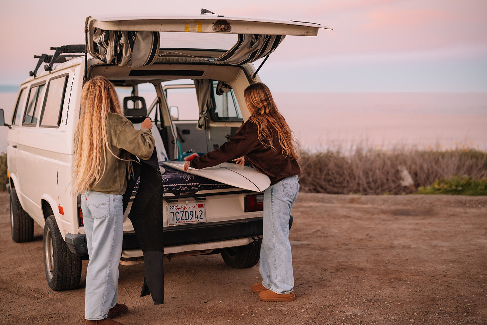 Two people unloading a surfboard from a van in a desert-like setting with a sunset sky.