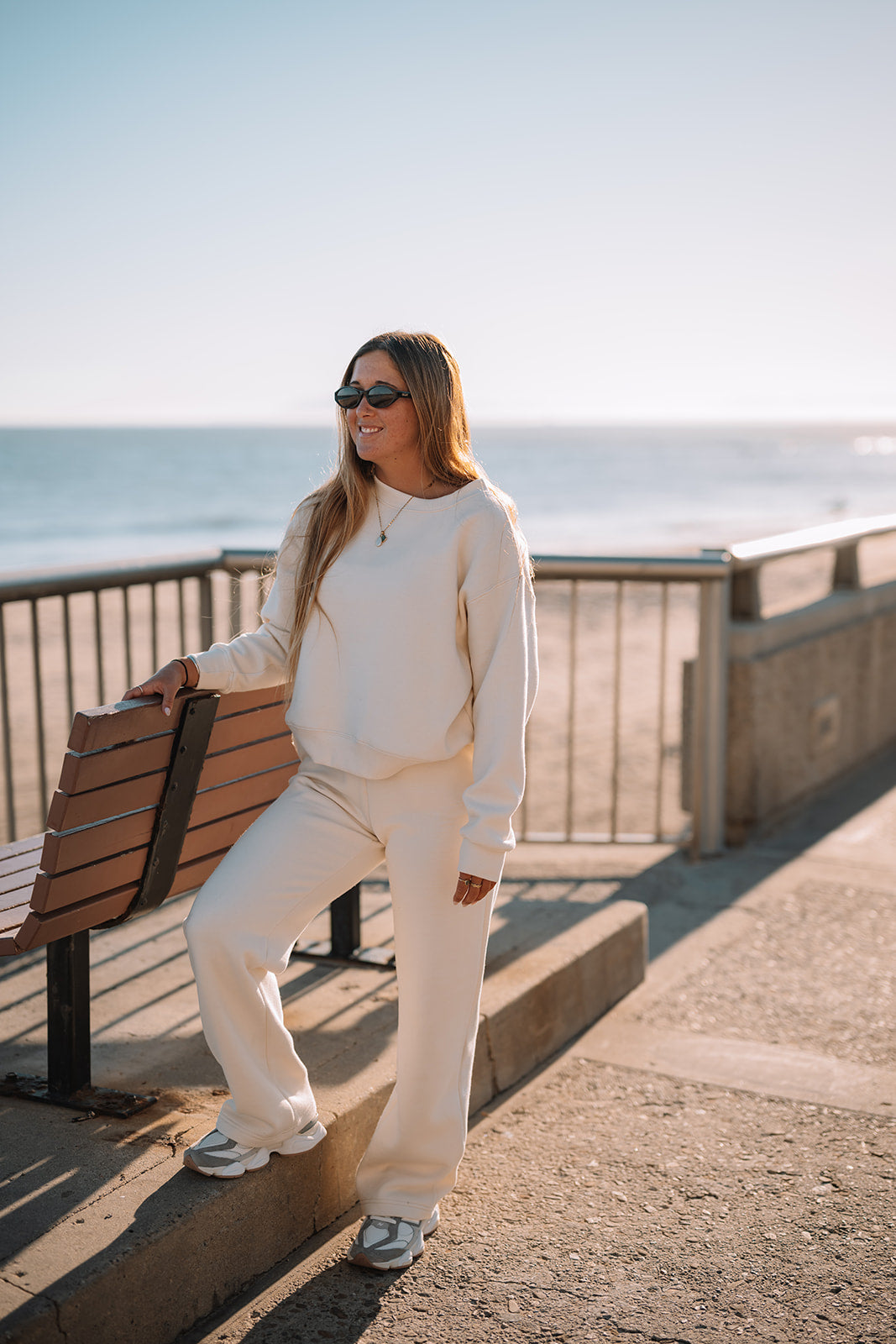 Woman in a white outfit standing near a bench by the ocean.