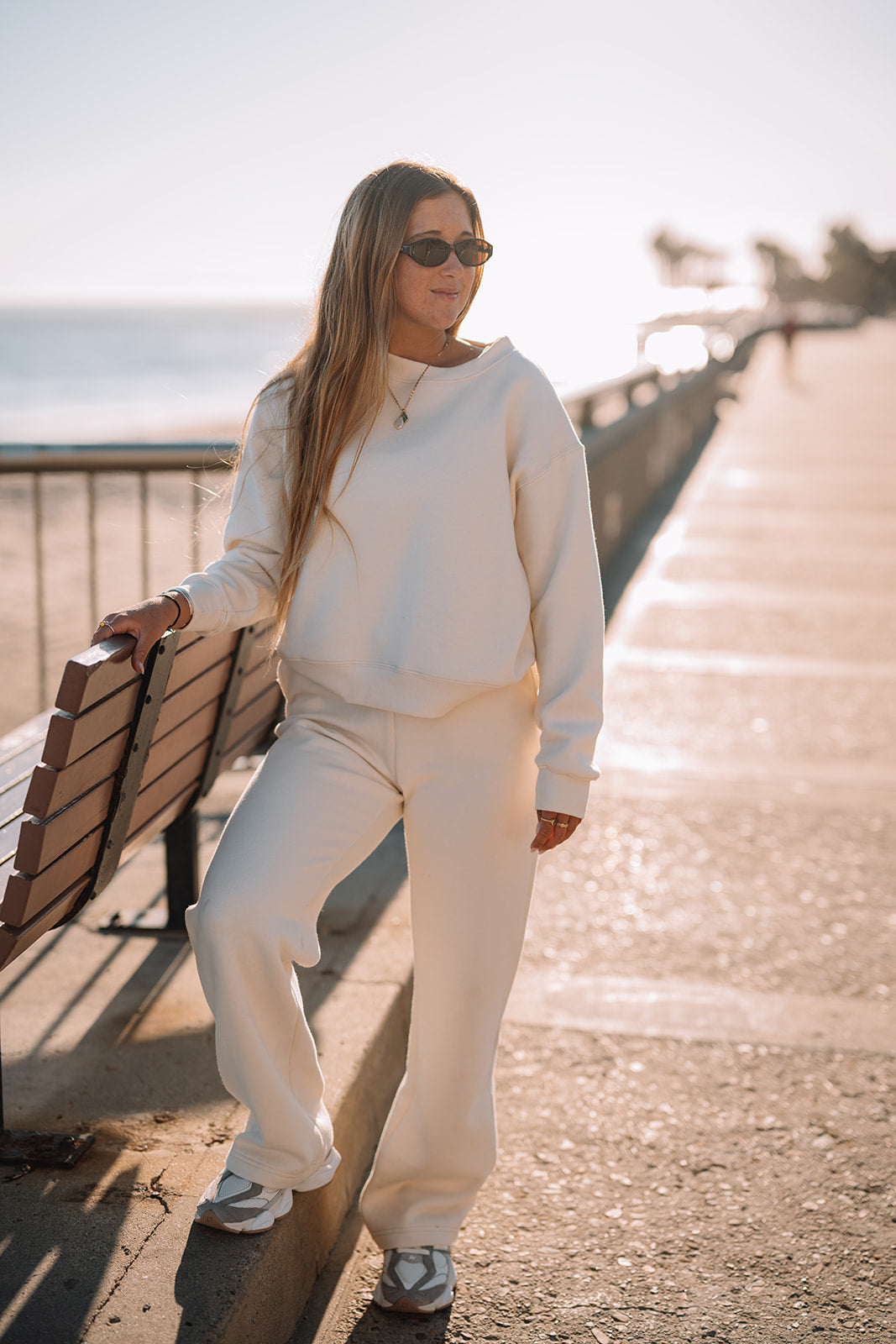 Person in a white outfit standing on a pathway with a bench and railing in the background.