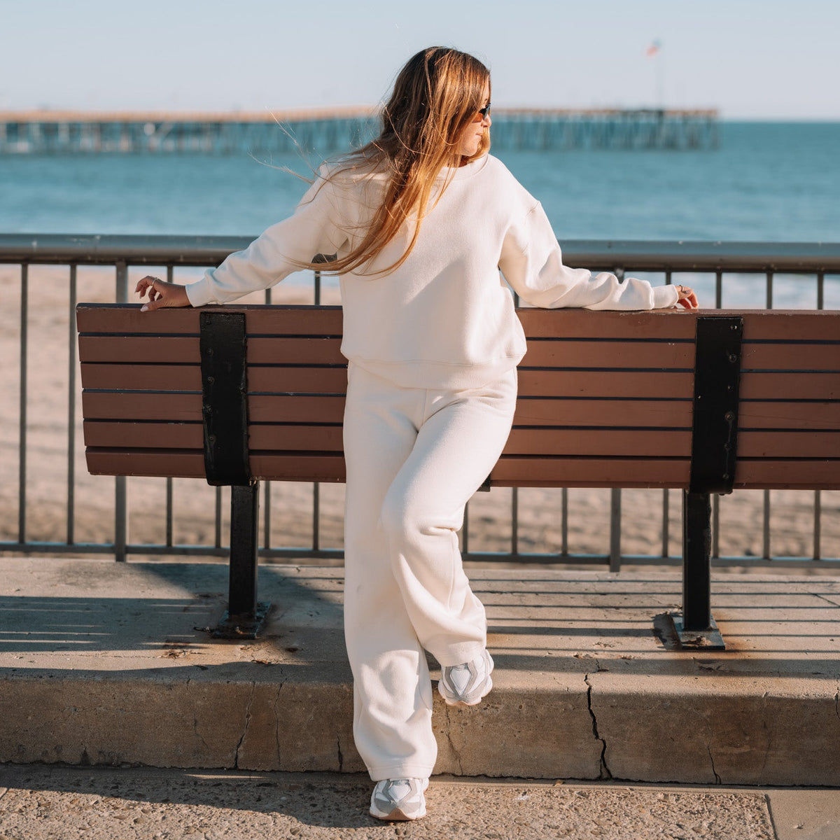 A woman in a white sweatshirt and matching sweatpants stands with her back to the camera, leaning on a wooden bench at a seaside location with the ocean and pier in the background.