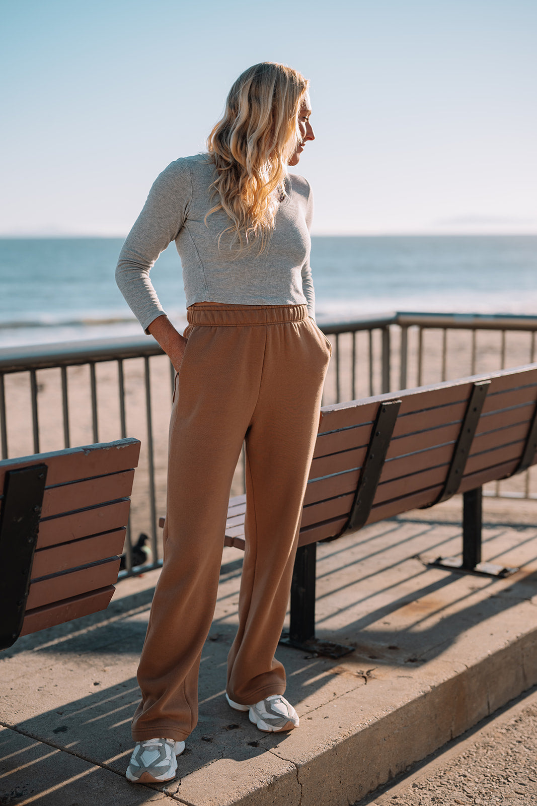 Woman standing on a boardwalk by the ocean wearing a gray top and brown pants.