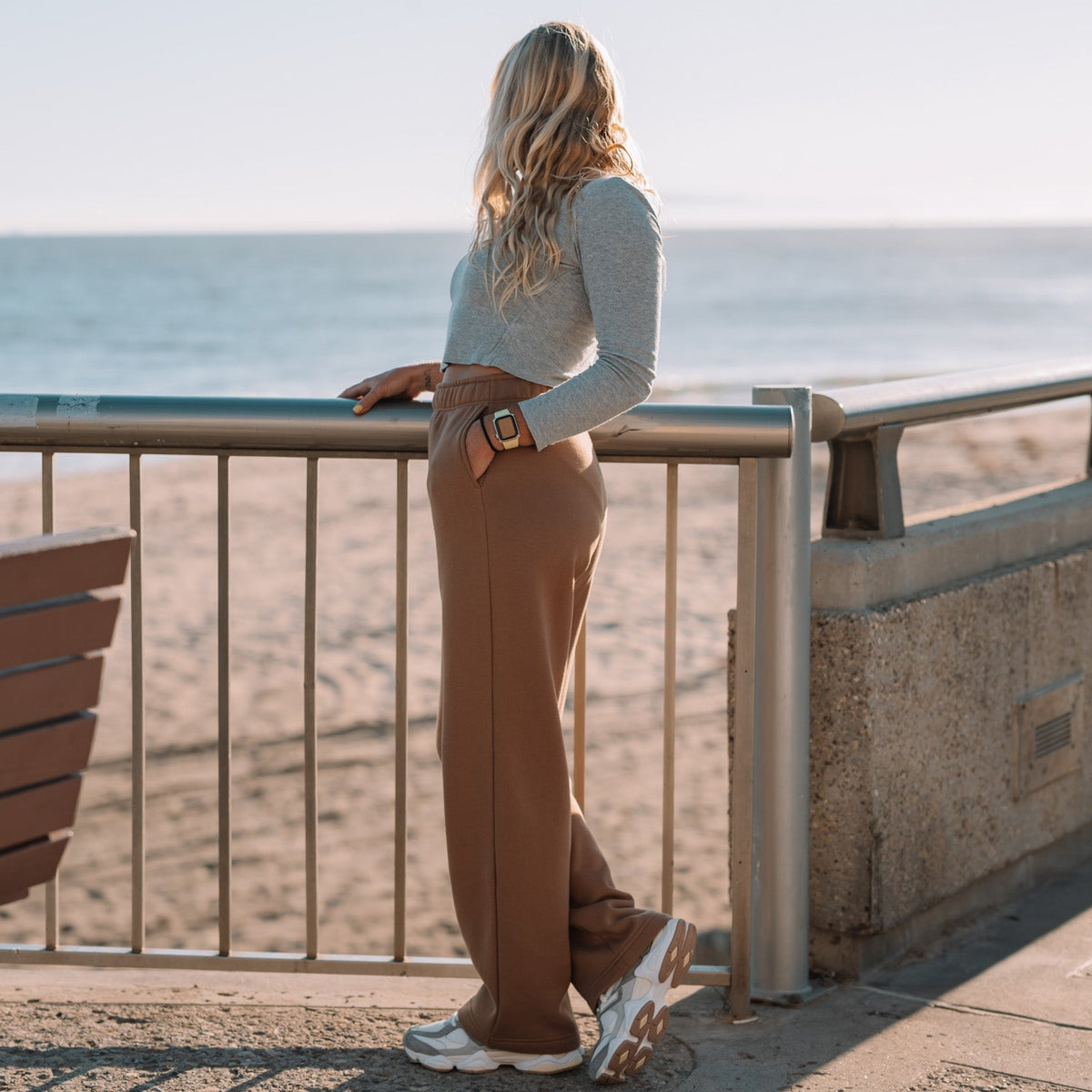 A woman stands on a seaside boardwalk railing, viewed from behind, wearing a light gray long-sleeve top, brown wide-leg pants, and white sneakers. The background shows the ocean, sand, and a concrete walkway.