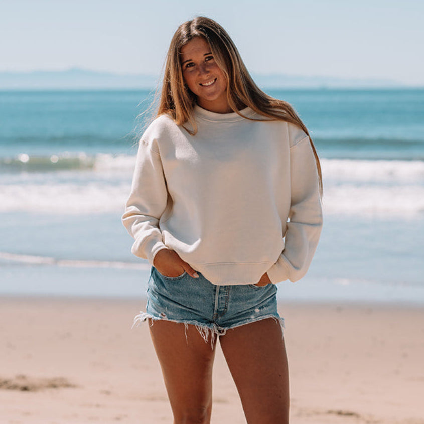 Woman standing on a beach wearing a white sweater and denim shorts with a clear blue sky and ocean in the background.