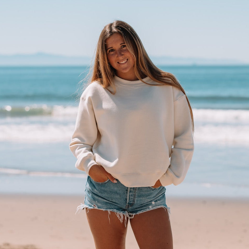 Woman standing on a beach wearing a beige sweater and denim shorts.