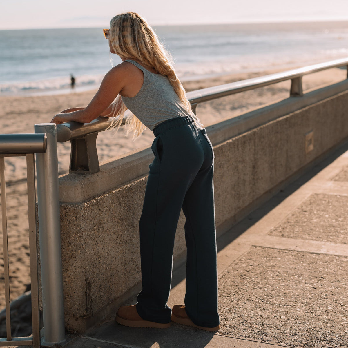 A woman with blonde hair stands on a seaside boardwalk railing, looking out toward the ocean at sunset. She is wearing a gray sleeveless top and dark wide-leg pants.