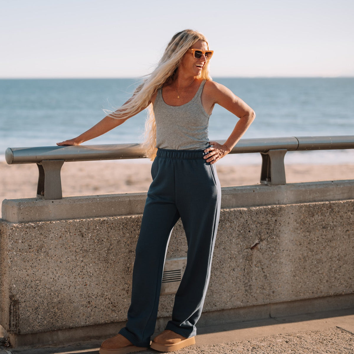 A woman with long blonde hair and sunglasses stands on a beach boardwalk next to the ocean, wearing a light gray ribbed tank top and dark gray wide-leg pants.