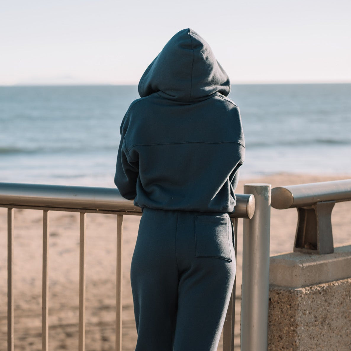 A person wearing a matching dark blue hoodie and sweatpants stands with their back to the camera, looking out over a beach railing at the ocean.