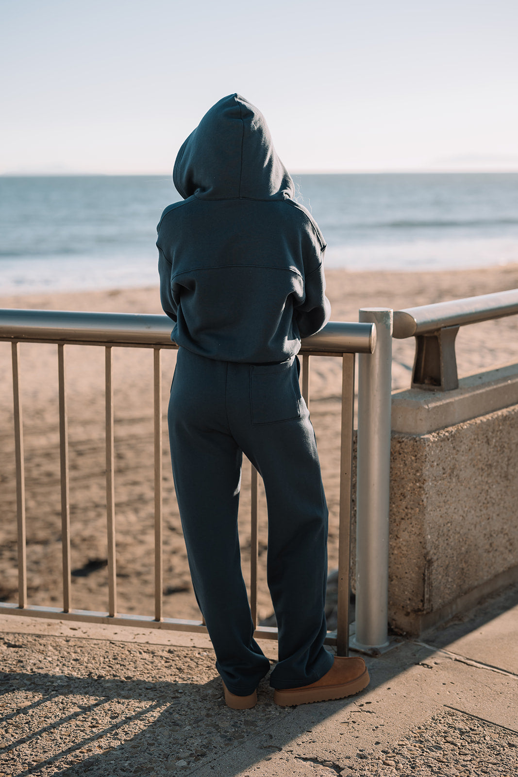 Person wearing a blue hoodie and pants standing by a railing with a beach and ocean view.