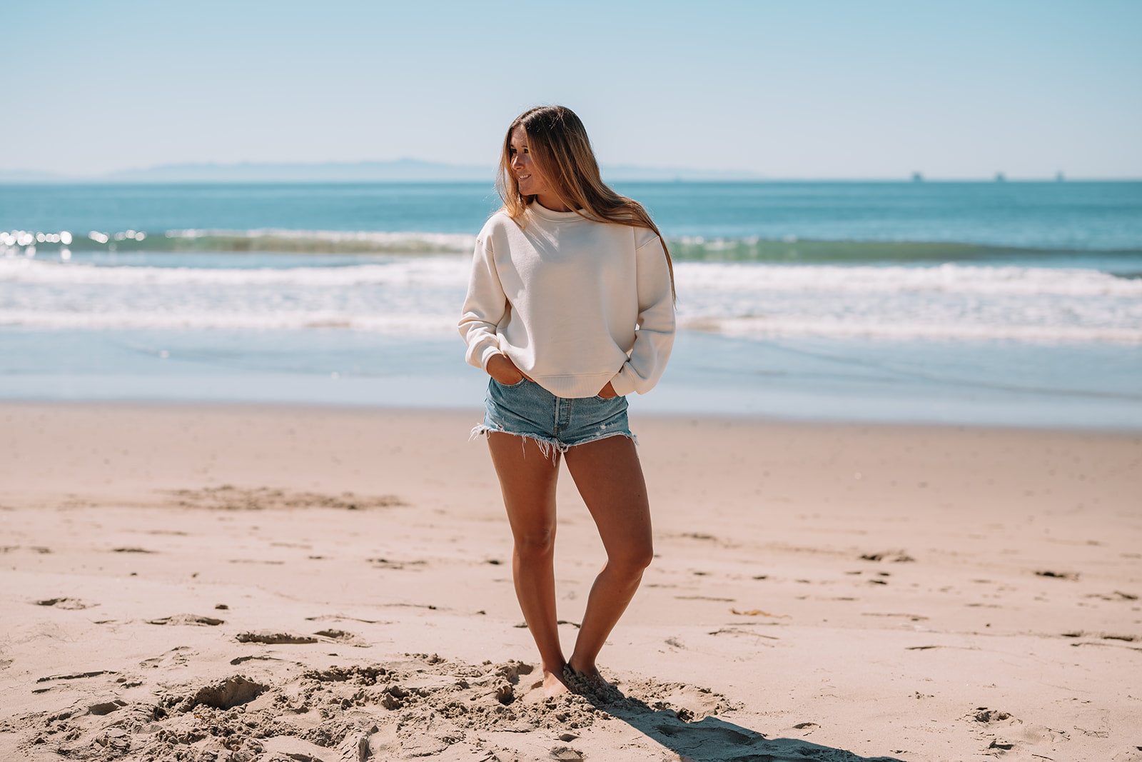 Woman in white sweatshirt and denim shorts standing on a sandy beach with ocean waves in the background