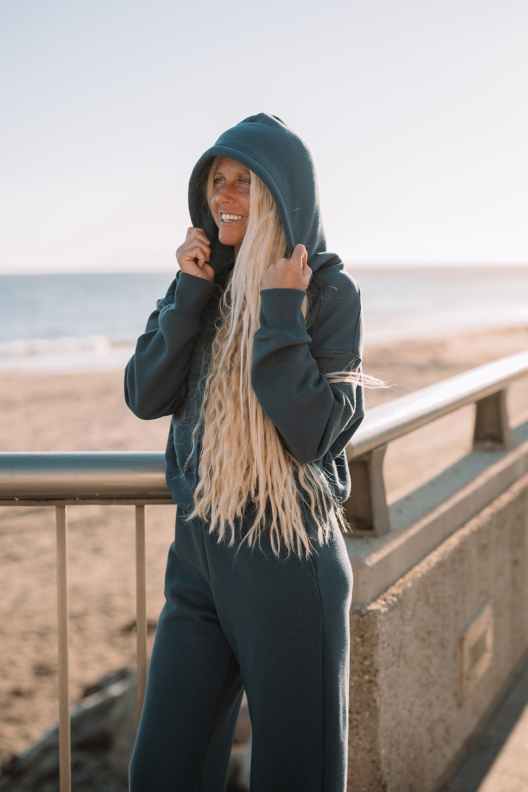 Person wearing a dark hoodie and pants standing by a railing with a beach in the background