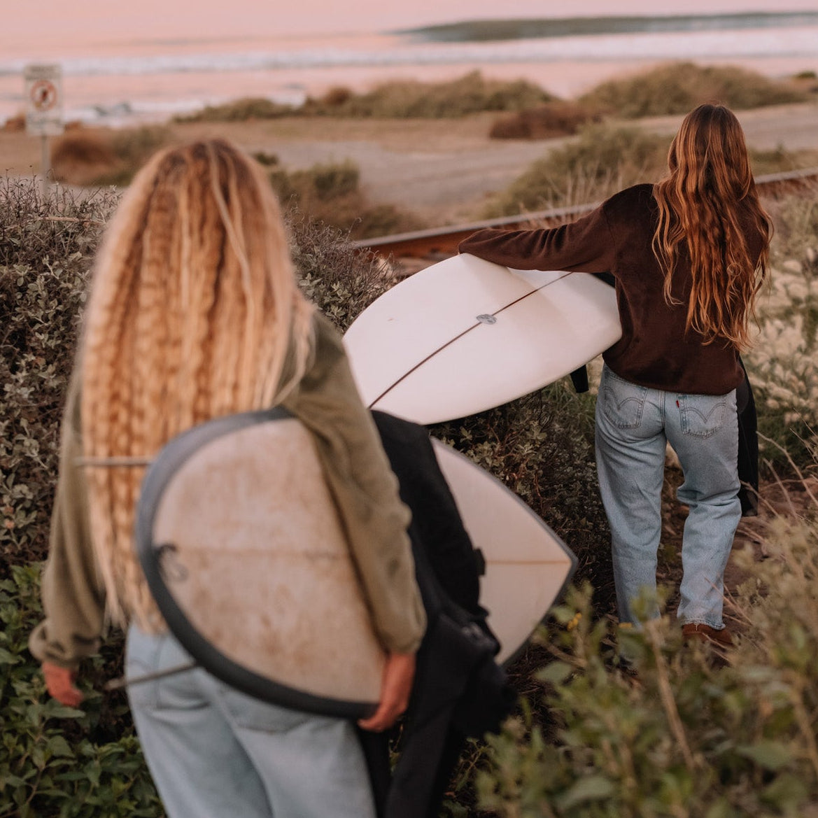 Two women carrying surfboards walk along a grassy path near railroad tracks at dusk, with a pink-toned ocean and distant hills in the background.