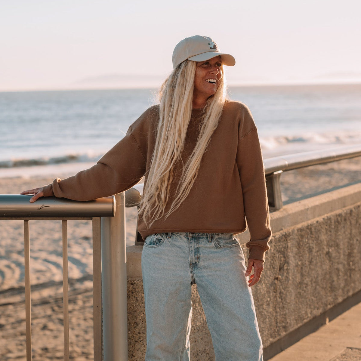 A woman with long blonde hair stands on a beach boardwalk at sunset, leaning on a metal railing. She is wearing a brown long-sleeve top, light blue wide-leg jeans, and a beige baseball cap.
