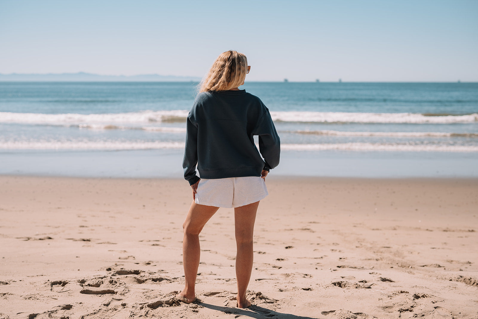 Person wearing blue crewneck and standing on a beach looking at the ocean 