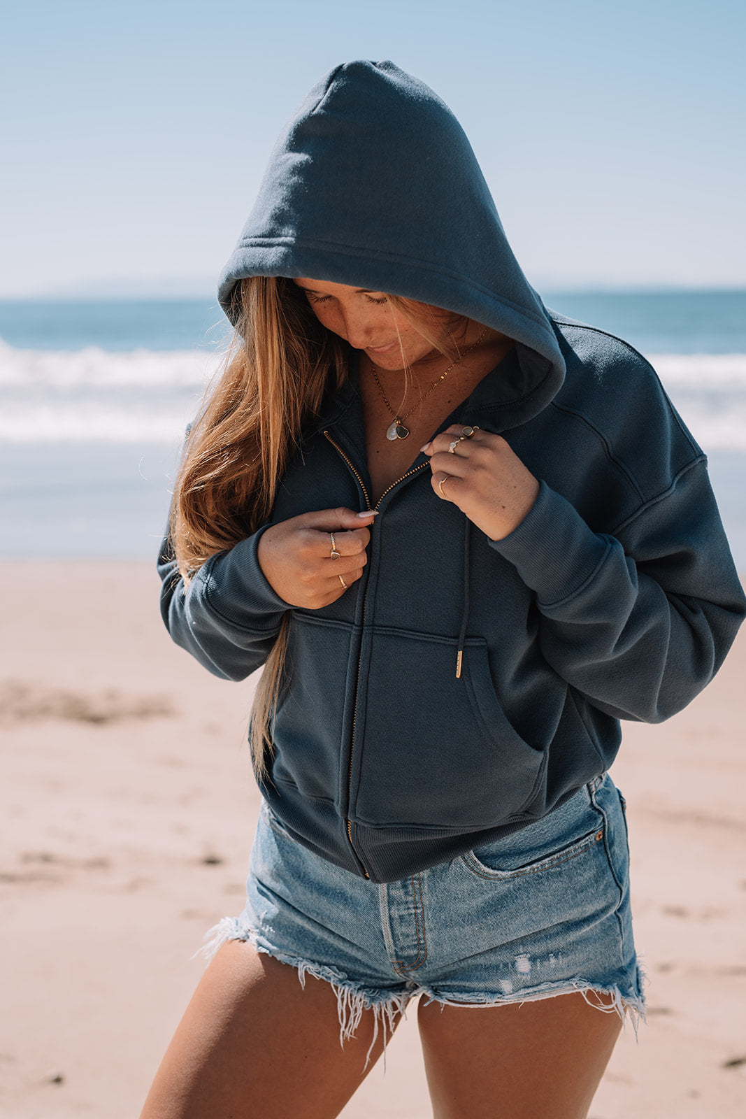 Girl unzipping blue hoodie with ocean in the background.
