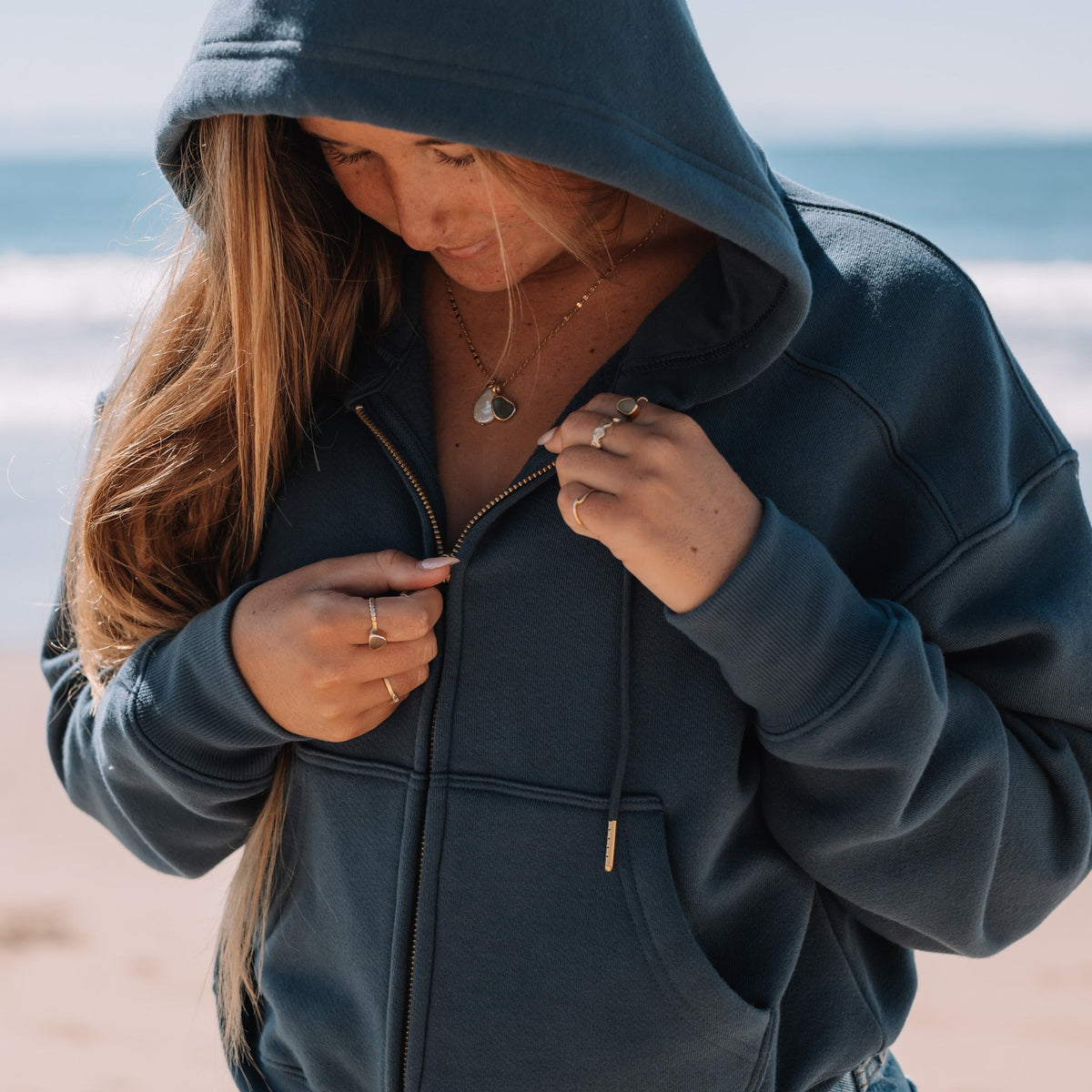 A woman wearing a navy blue zip-up hoodie with a drawstring hood, standing on a sandy beach with the ocean in the background. She is looking down while adjusting the zipper.