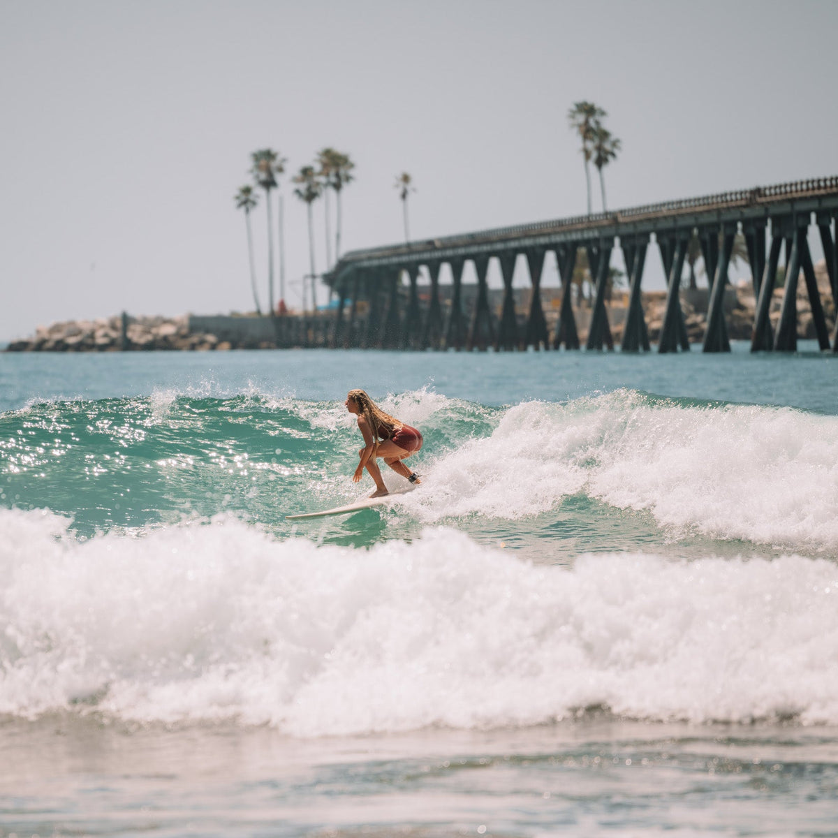 Person surfing in the ocean with a pier and palm trees in the background