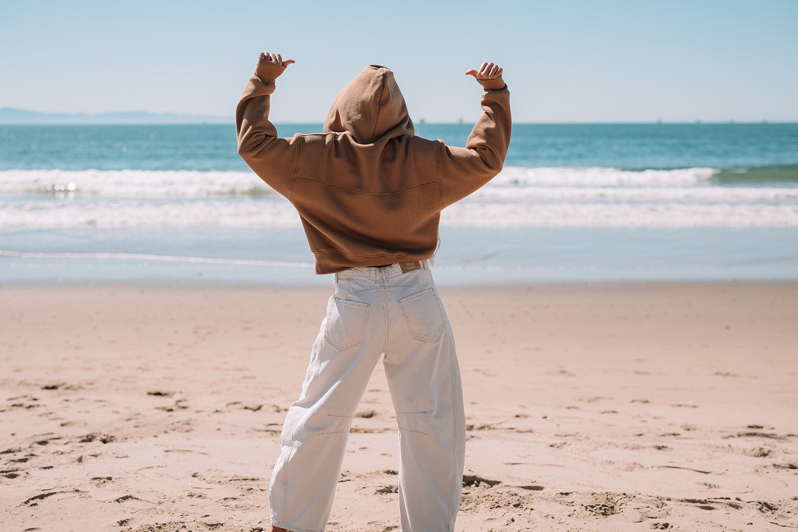 Person wearing a brown hoodie and white pants standing on a beach with arms raised towards the sky.