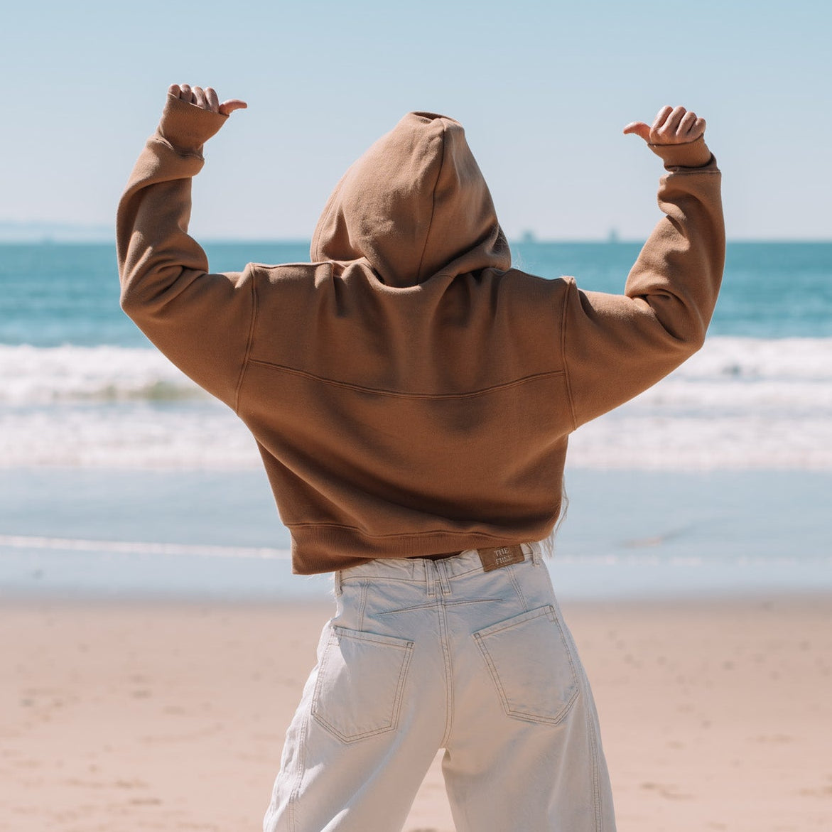 Person wearing a brown hoodie and white pants standing on a beach with arms raised.