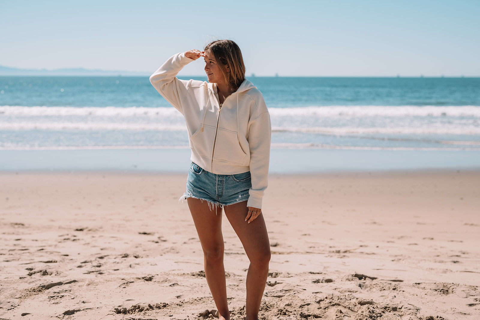 Woman standing on a beach wearing a white hoodie and denim shorts, with ocean waves in the background.