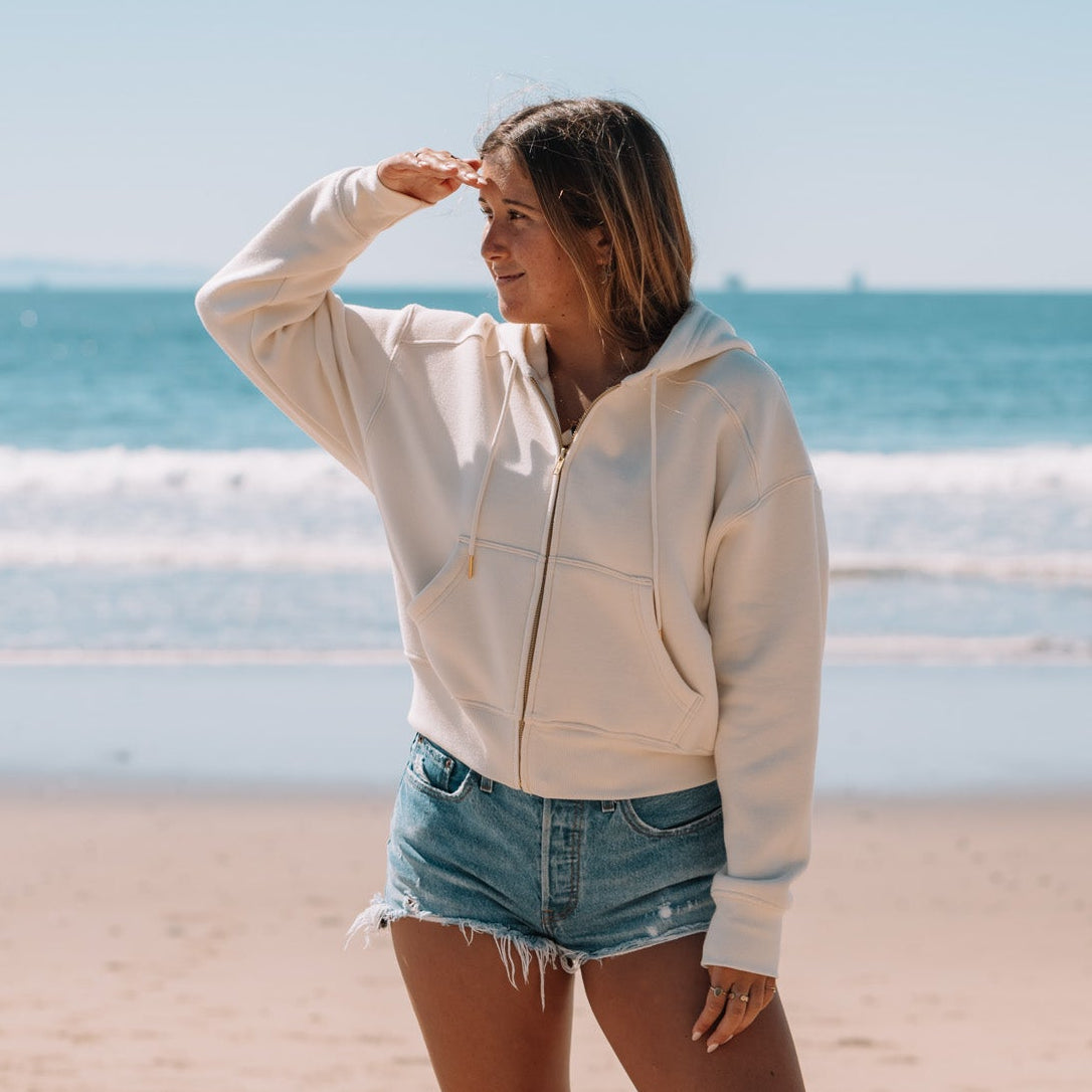 Woman standing on a beach wearing a white sweatshirt and denim shorts, with ocean waves in the background.