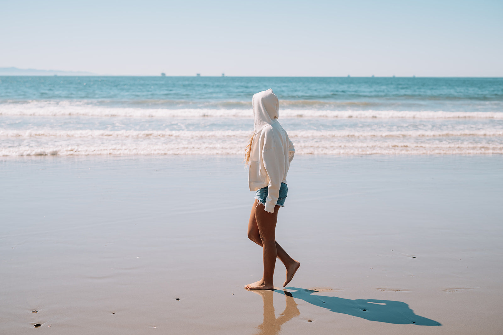Person in a white hoodie walking on a beach with clear blue sky and ocean.