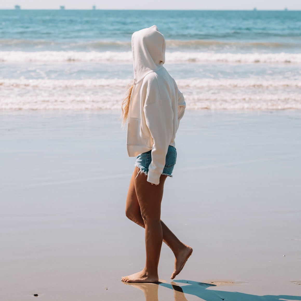 Person walking on a beach wearing a white hoodie and shorts