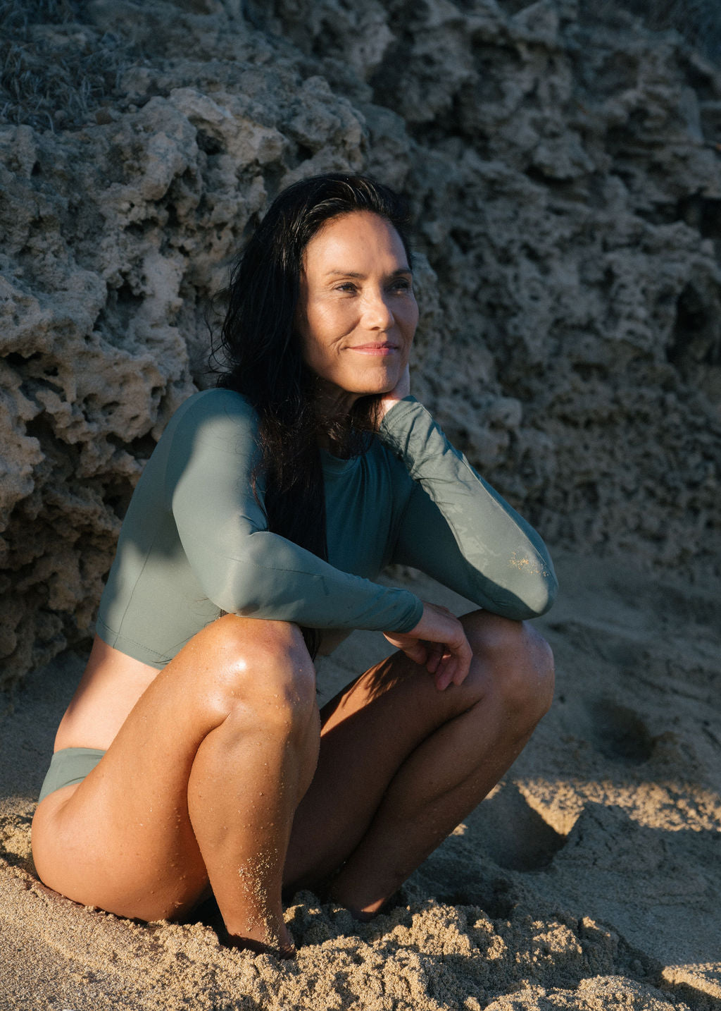 Woman in a green swimsuit sitting on a sandy beach with textured rock wall in the background