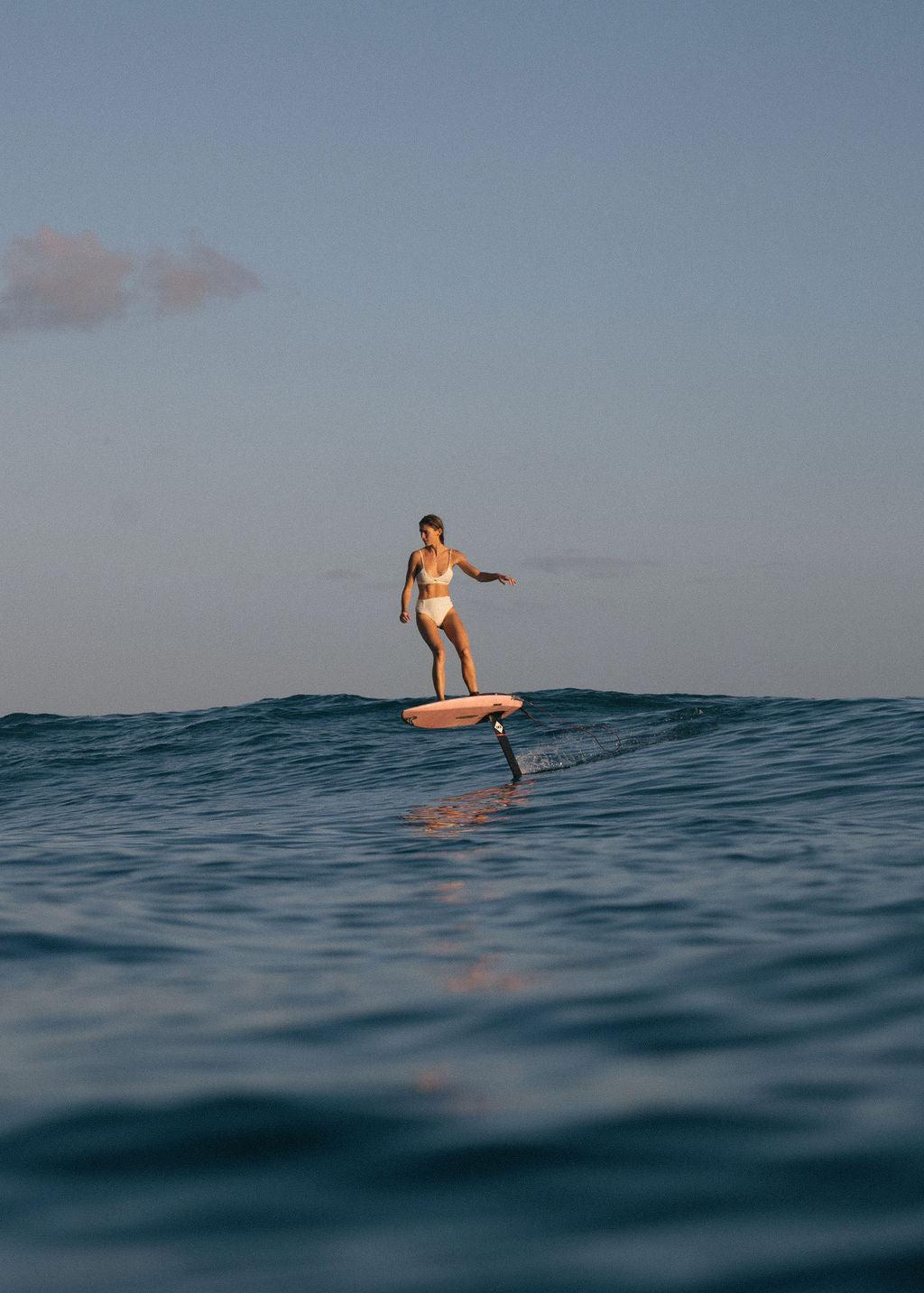 Female athlete in white bikini riding a pink hydrofoil board.  She is turning on a wave.