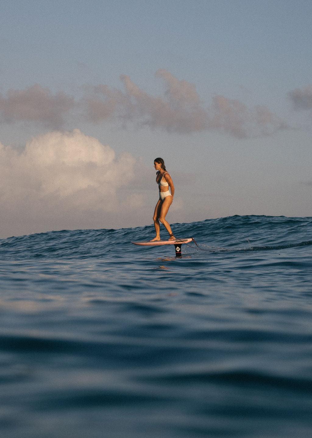 Female athlete riding a hydrofoil board in the ocean.  She is wearing a white bikini and the sun is shining on her.