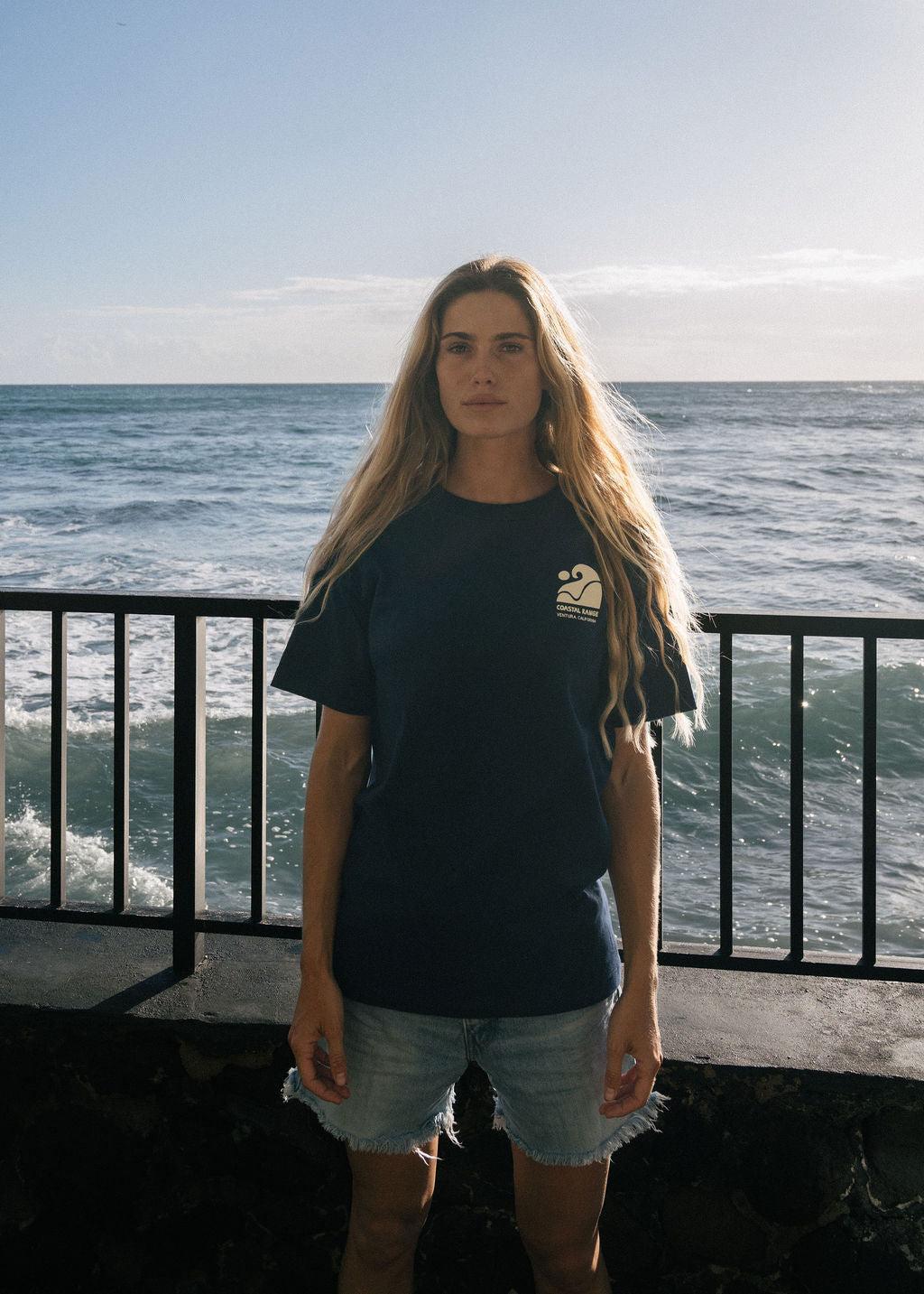 female with blue t-shirt and cutoff jean shorts standing in front of ocean