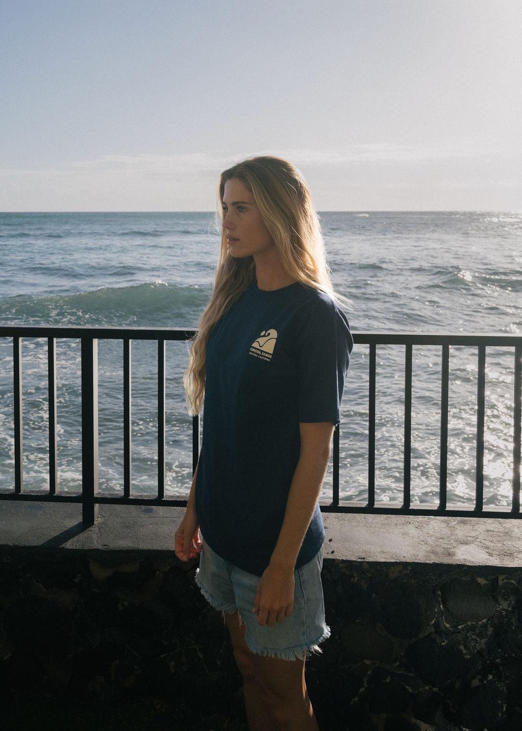 girl with long hair, wearing blue t-shirt and cut-off jean shorts looks toward the beach