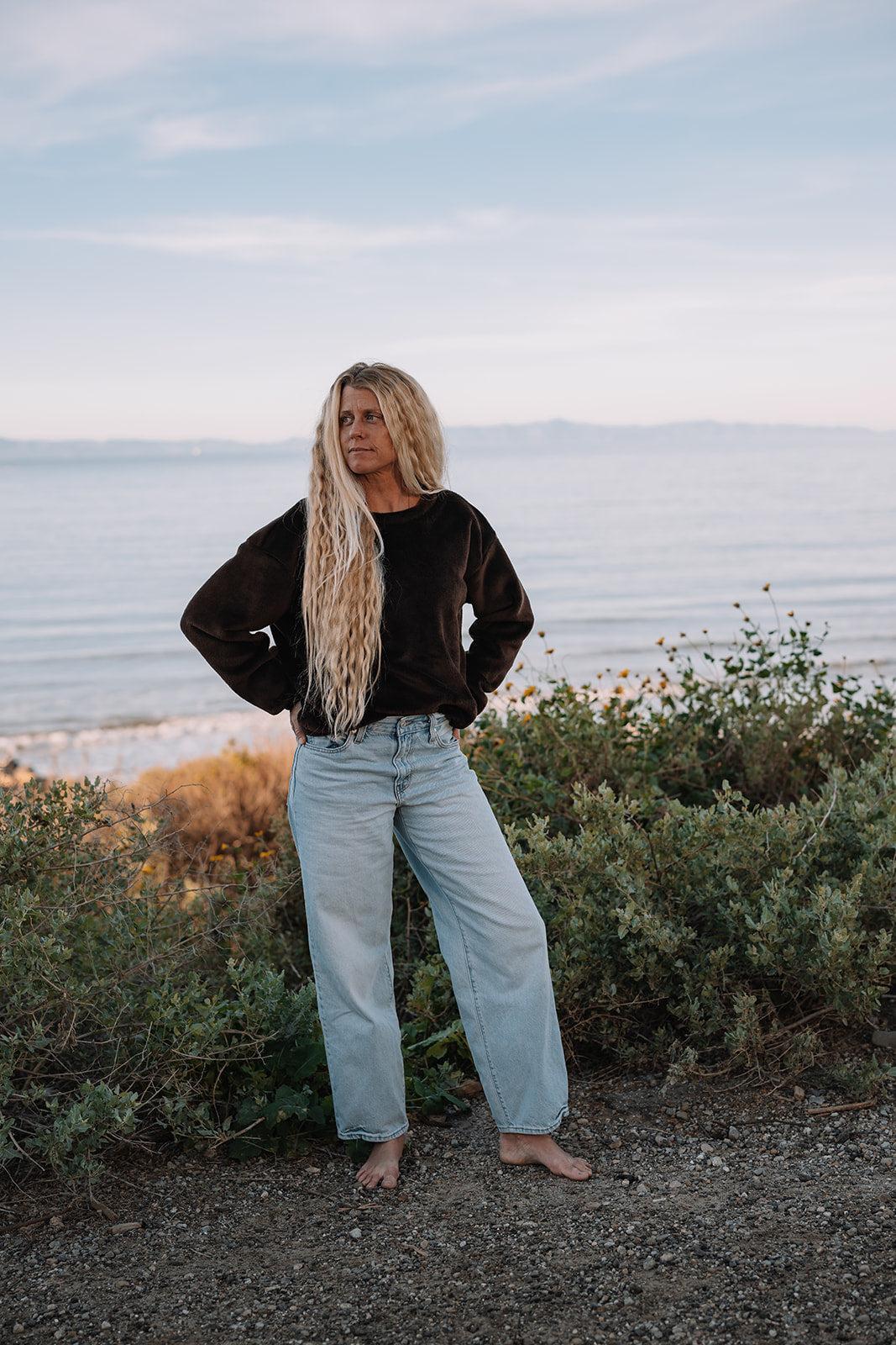 Woman in a dark brown Harborview Fleece Sweater and light wash jeans by the ocean.