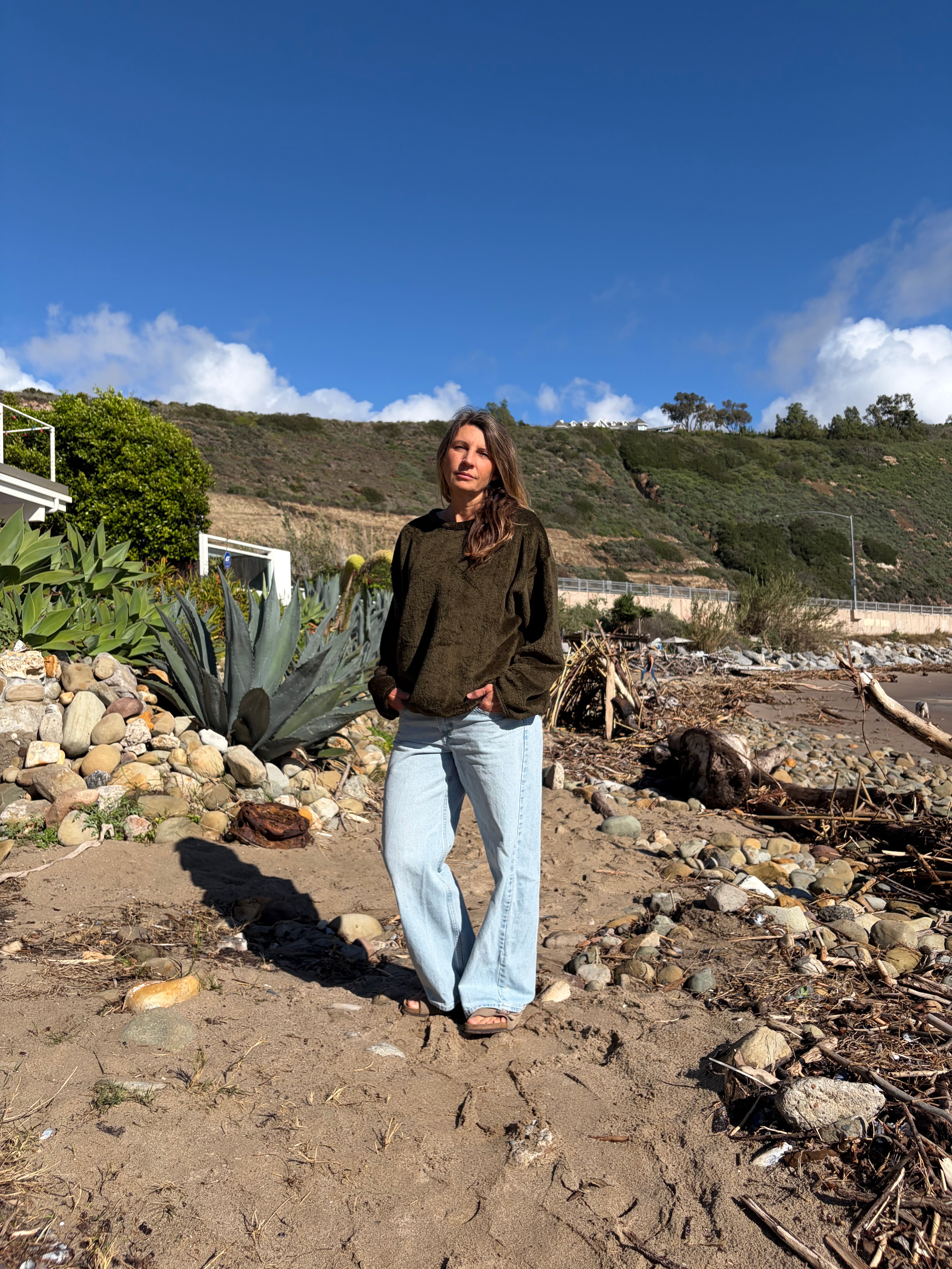 Woman on a beach wearing a relaxed-fit, dark olive green fleece sweater with light wash jeans.