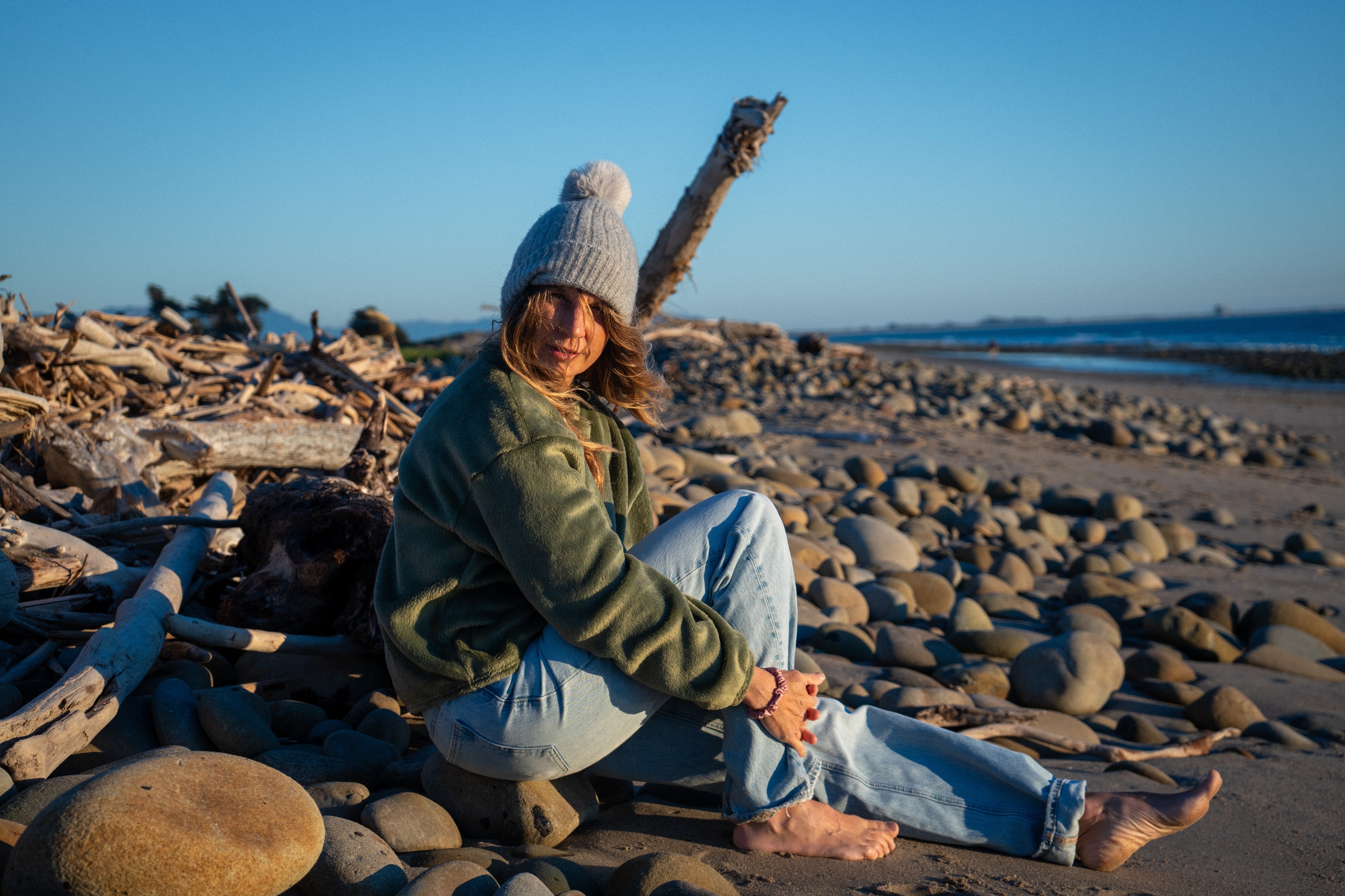 Woman in a relaxed green fleece sweater and grey pom-pom beanie sitting on a rocky beach.