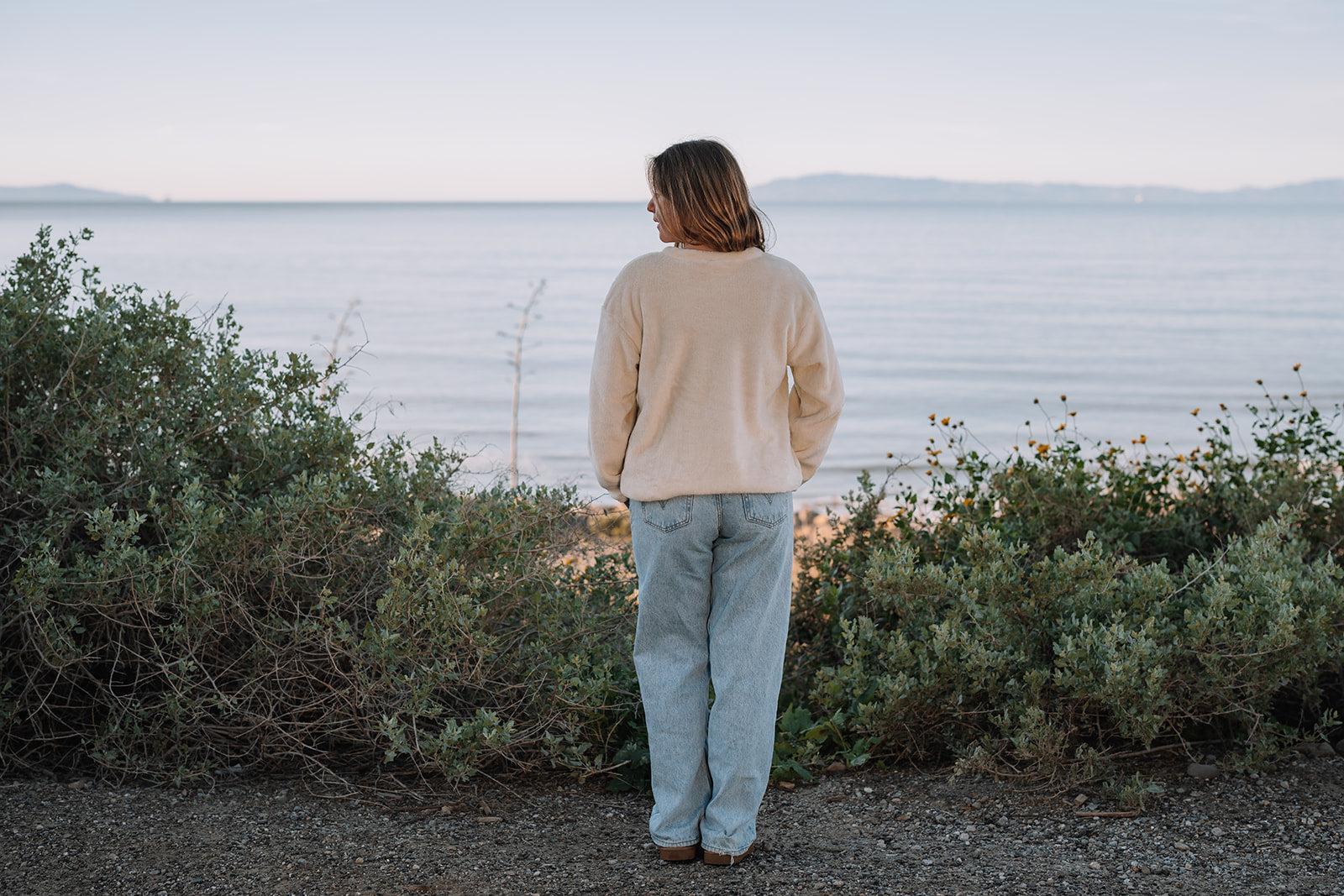 Woman in a relaxed-fit, cream Harborview Fleece Sweater and jeans, facing a calm ocean at sunset.