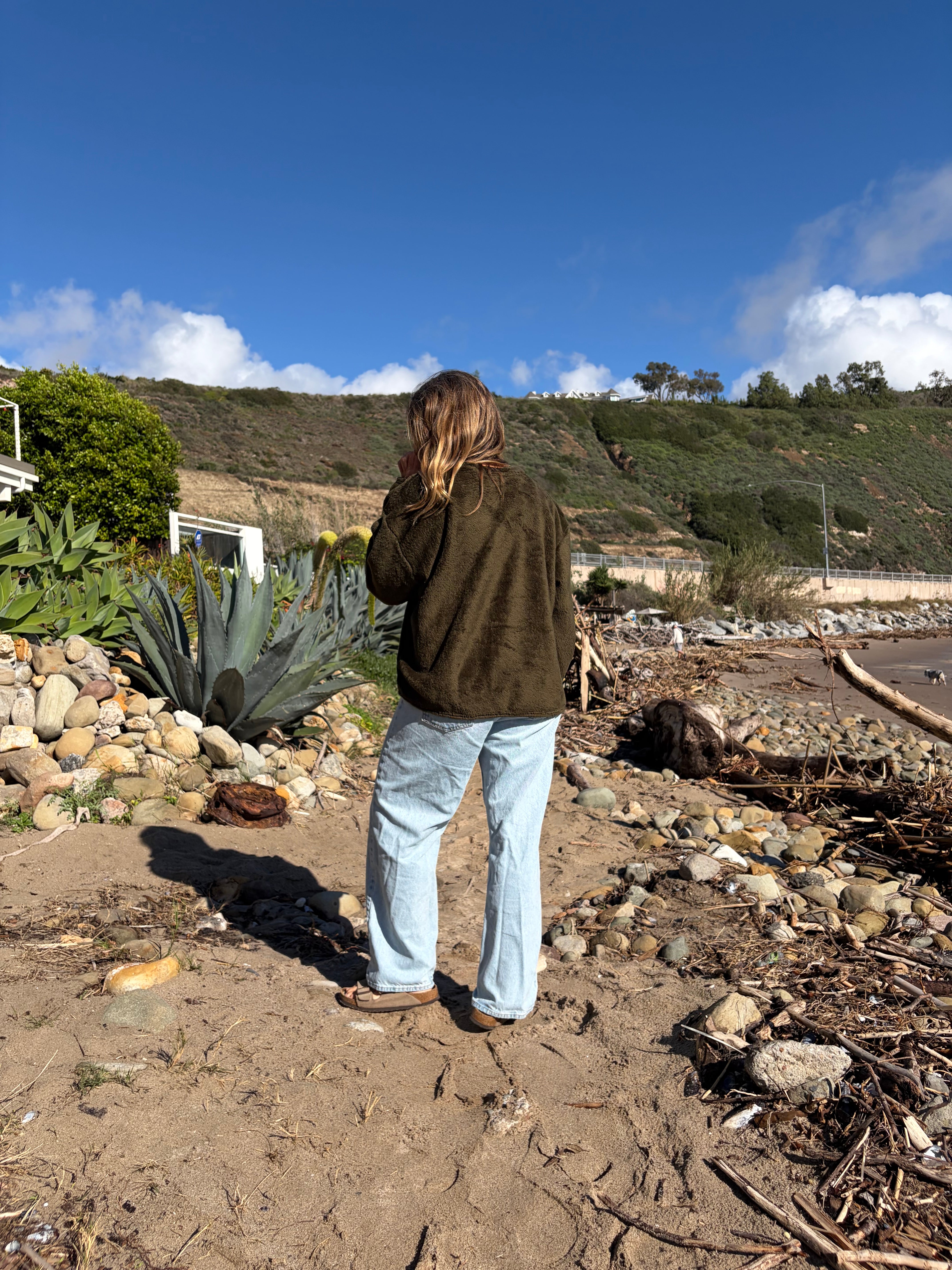 Woman in a relaxed-fit olive green fleece sweater and light wash jeans on a sandy beach.