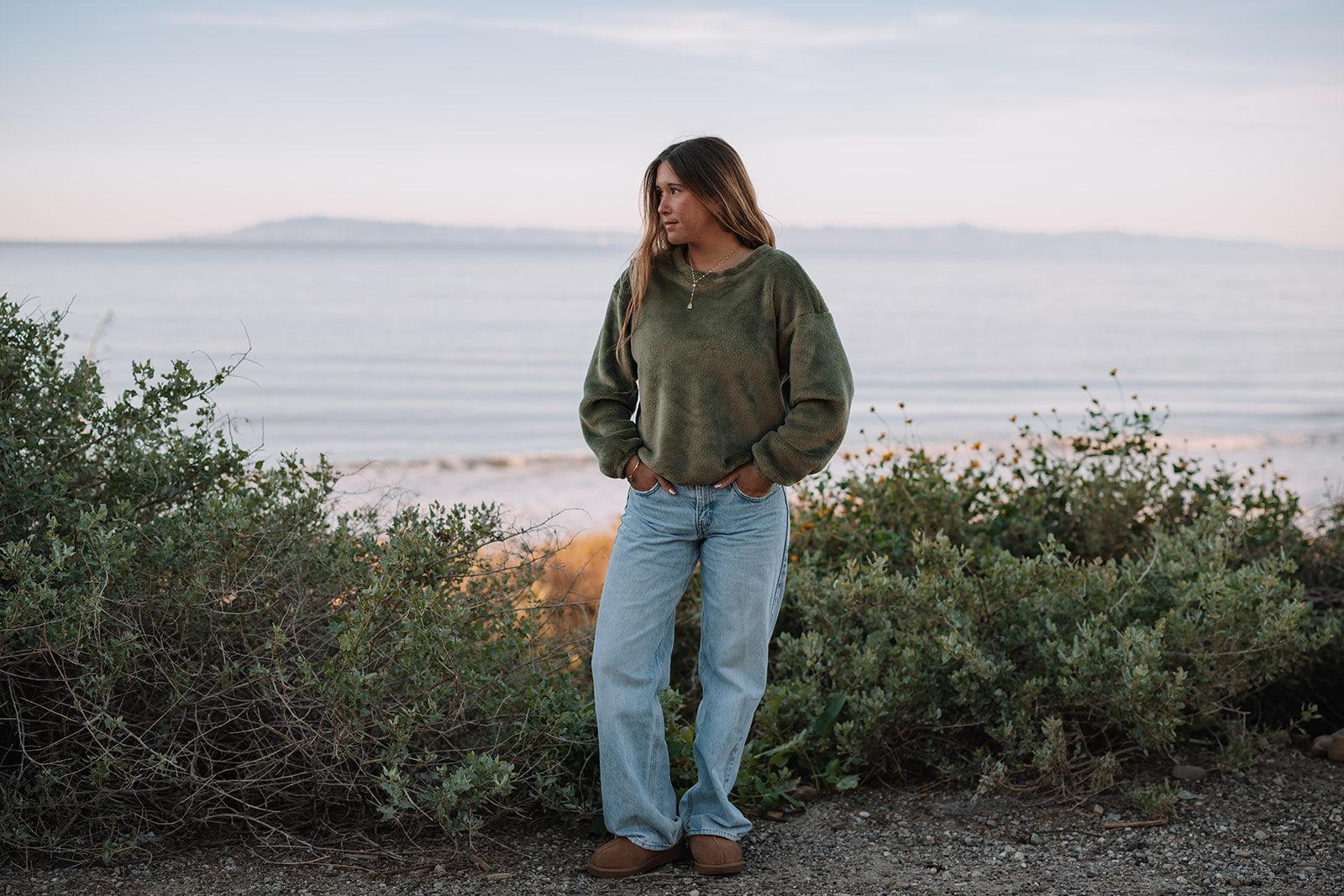 Woman in a plush olive green Harborview Fleece Sweater and light wash jeans by the ocean.