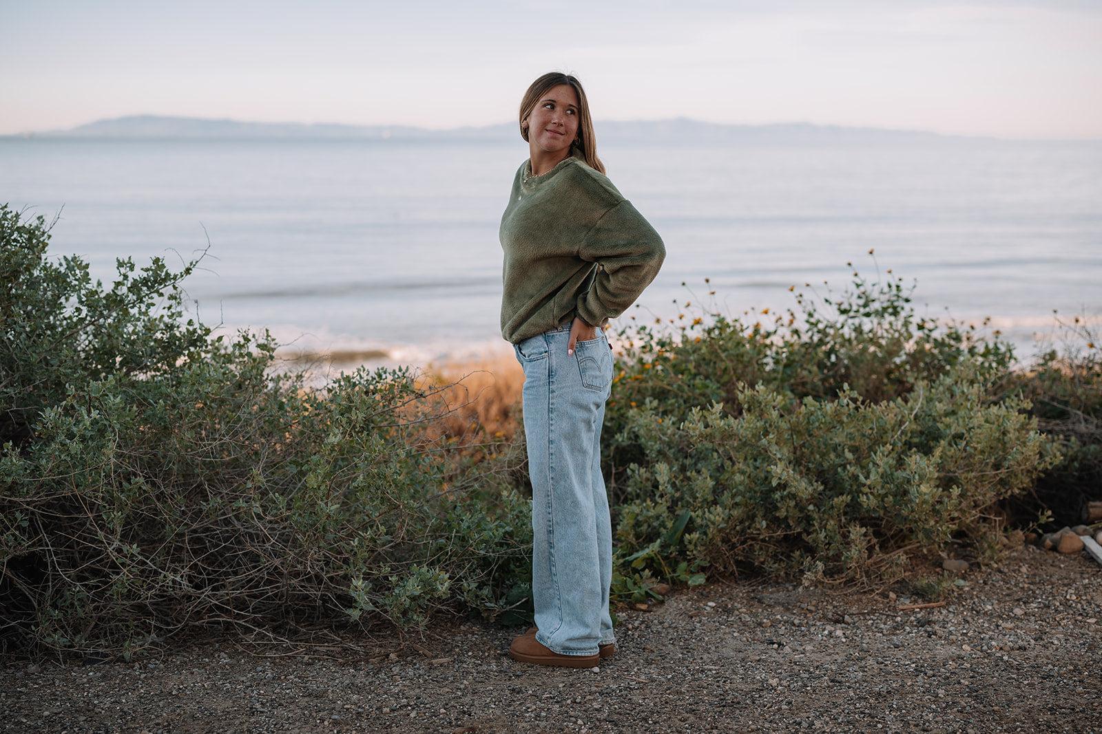 Woman in a relaxed-fit olive green fleece sweater and light-wash jeans by the ocean.