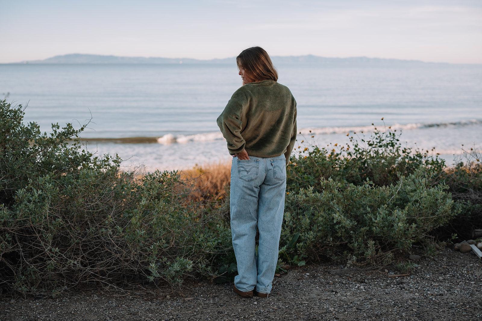 Woman in an olive green Harborview Fleece Sweater and light wash jeans, facing a calm ocean.
